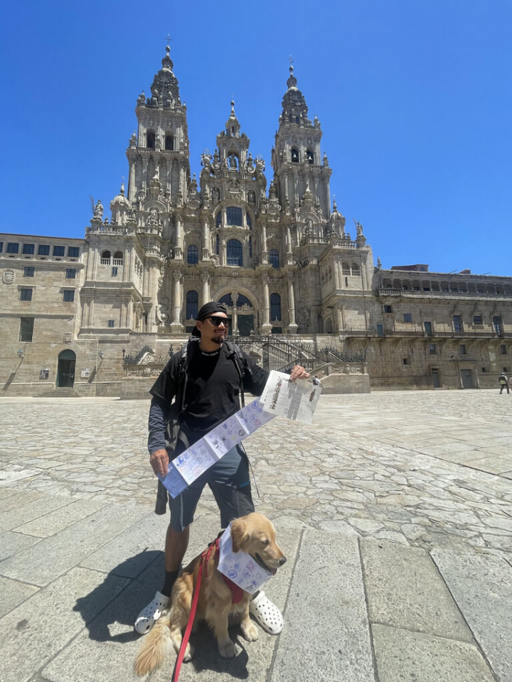 來自台灣的黃金獵犬Money和主人Burger Han（臉書帳號），徒步完成12天共315公里的朝聖之行後，在終點站聖地牙哥德孔波斯特拉主教座堂前，與蓋滿沿途紀念戳印的朝聖者護照合影。（Burger Han提供）