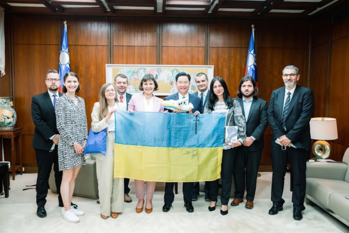 外交部長吳釗燮與訪團乙行及攜自俄烏戰事前線的烏國國旗合影。（左起：捷克駐台代表史坦格、烏籍EVC研究員馬卡羅維奇、烏籍EVC研究員莉巴邱、People in Need執行長帕納德、烏克蘭反貪腐行動中心執行長卡雷紐克、吳部長、EVC執行長揚達、烏克蘭國際勝利中心主席霍普科、EVC台北辦事處負責人葉浩勤、People in Need共同創辦人布拉澤維奇。）