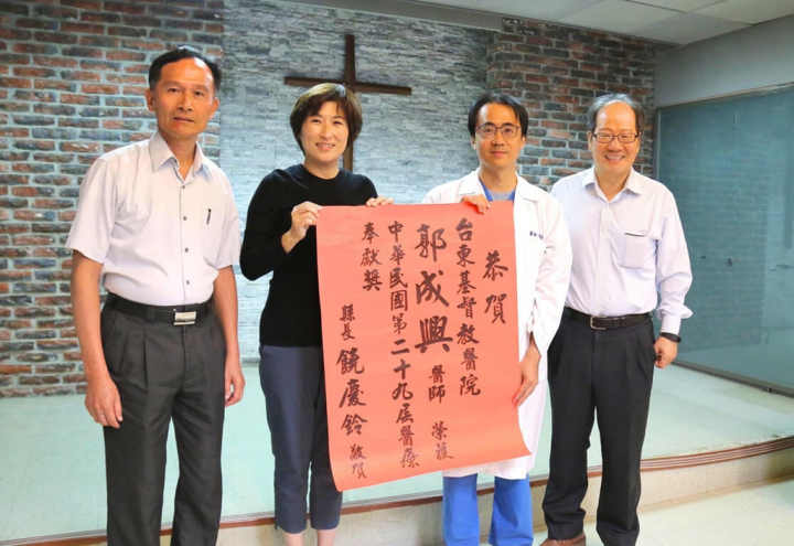 高雄醫學大學學士後醫學系校友郭成興（右2）毅然從繁華故鄉台北市遠赴台東服務，2019年榮獲第29屆「醫療奉獻獎」，台東縣長饒慶鈴（左2）貼紅榜祝賀。（高醫提供）