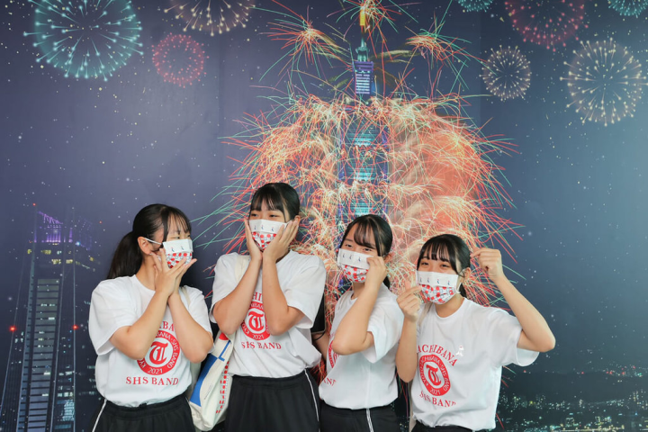 日本京都橘高校吹奏部應邀來台參與國慶演出，也把握空檔時間參訪台北101，學生們興奮在101大樓看板前合影留念。（中華文化總會提供）