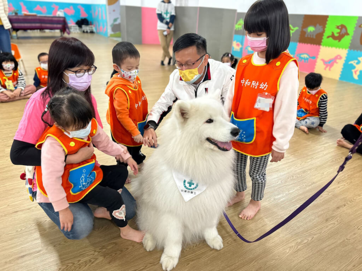 新北市中和國小附設幼兒園30日結合主題教學課程「聽我說，我愛你」，邀請台灣狗醫生協會的狗醫生「魯蛋」（前中）到幼兒園參與教學，新北市教育局長張明文（右2白衣者）也到場，陪孩子為狗醫生梳毛等。（新北市教育局提供）