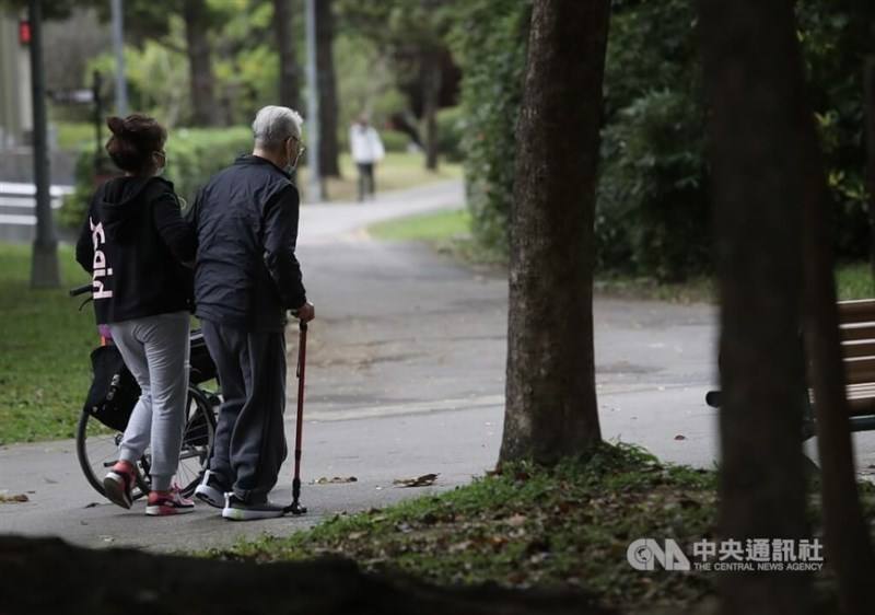 An elderly man (right) on a cane and his caregiver take a stroll at a park.