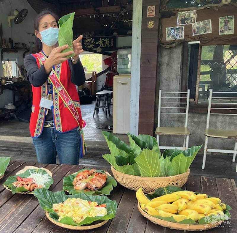 A woman presenting a leaf-wrapped meal. Inside is a soft rice and yam mixture with fresh onions and sausage pieces. Also on the table is stir-fried cabbage, roasted chicken, and bananas.