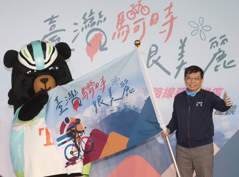 交通部長王國材16日在台北松山車站出席「台灣騎時很美麗」活動，出發前，台灣觀光吉祥物「喔熊組長」（左）授旗給王國材（右），祝福騎乘順利