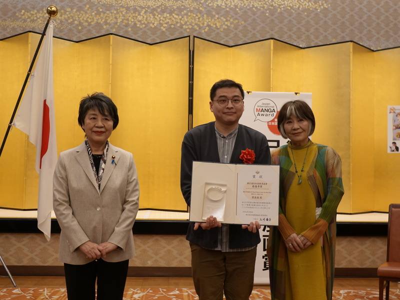 由日本外務省舉辦的第17屆日本國際漫畫賞5日晚間在東京舉頒獎典禮，台灣漫畫家簡嘉誠（中）以作品「青空下的追風少年」獲最優秀獎（金獎），從日本外務大臣上川陽子（左）手中獲頒獎狀、從評審委員長里中滿智子（右）手中接下獎盃。