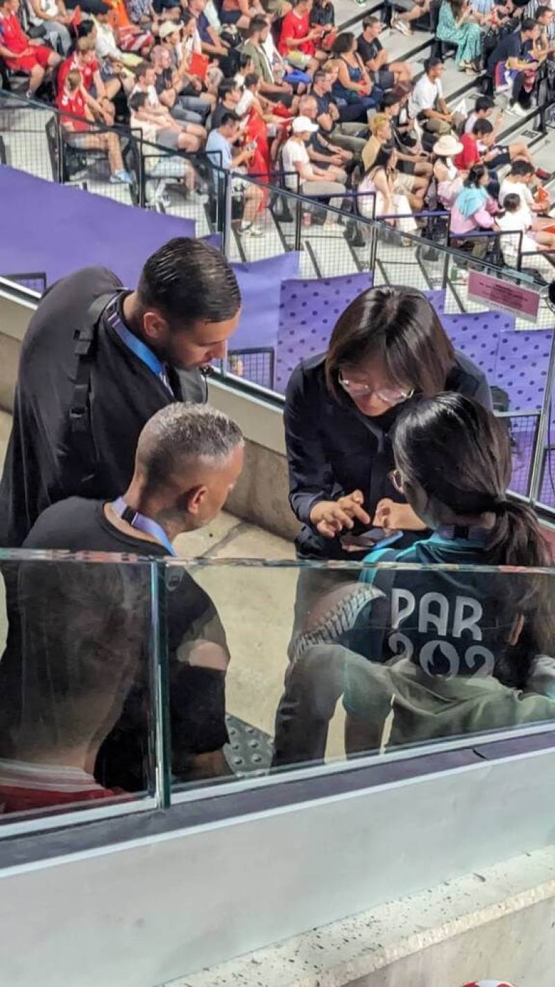 A woman (right, holding a mobile phone) shows her phone to staff by the court of the gold medal match of the badminton men's doubles on Sunday at the Paris Olympics. She was suspected to be pointing out fans who carried flags and banners to be confisticat