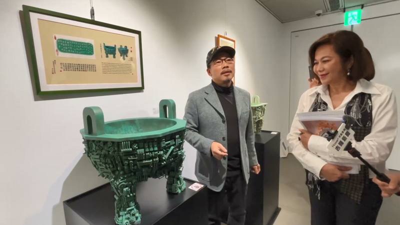 作詞人方文山（左）創作仿青銅器毛公鼎，外觀以各式機械構造呈現，鼎內「鐘鼎文」則取用由他作詞、歌手周杰倫演唱的「青花瓷」歌詞。基隆市副市長邱佩琳（前右）27日在開展記者會仔細聆聽方文山介紹作品。