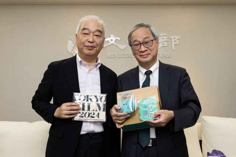 文化部長李遠（右）致贈「臺灣文化in大阪．關西世博」吉祥物「a-We」痛包及徽章等文宣品予東京影展資深策展人石坂健治（左），並說明「a-We」多元、包容、韌性的形象。