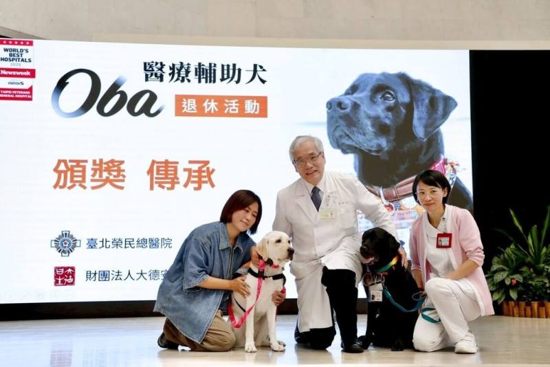 全台首隻駐點醫院的輔助治療犬「歐巴」（黑犬）16日在北榮院長陳威明（中）見證下正式退休，牠的工作將由3歲「學妹」歐佳（白犬）接手。（北榮提供）