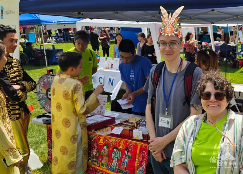 戴著臺灣原住民頭飾的中級班學生 Scot Harrington（右二）與初級班學生 Lydia Parenteau（右一）在文化攤位前與部分與會嘉賓志工合影