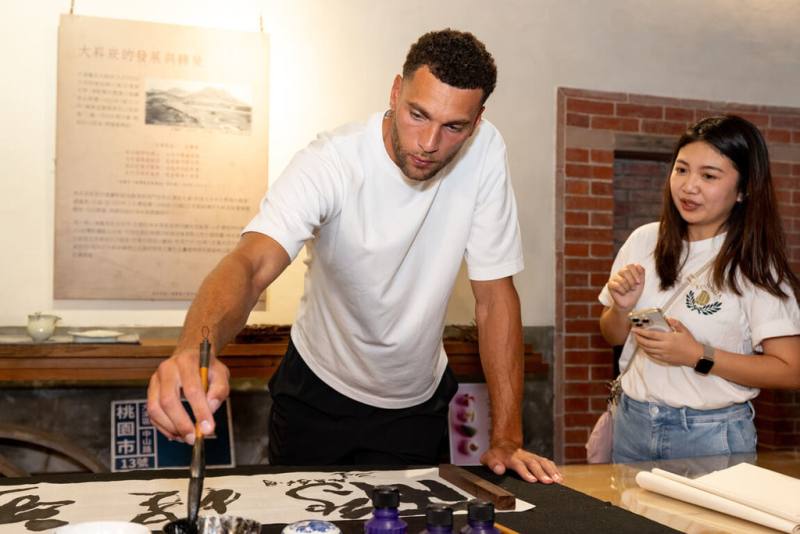 美國職籃NBA球星拉文（Zach LaVine）（左）2日走訪大溪老街，觀賞書法老師揮毫「鵬程萬里」，呼應他2度奪得NBA灌籃大賽冠軍的飛人形象，拉文親自在作品落款，也提筆寫下草書「飛」字留念。（桃園市政府觀光旅遊局提供）