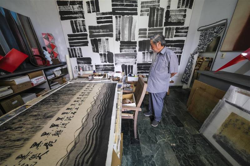 李再鈐在工作室內的身影。牆上為水墨作品〈爻〉（2024）的一部分；桌上則是他心繫社會，為花蓮土石流災民所繪製的新作。